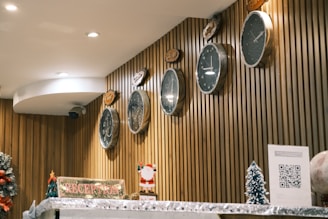Several clocks with different time zones are mounted on a wooden panel wall. A reception counter is adorned with festive decorations, including a small Santa figurine and miniature Christmas trees. A sign reading 'RECEPTION' is prominently displayed alongside a QR code on a piece of paper.