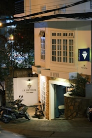 A cozy street-side café called 'Le Retour' with a small outdoor seating area consisting of a round table and two chairs. A scooter is parked nearby on the pavement beside a stone wall. The café features a combination of white and brick walls with large windows. The sign on the wall includes 'Vin et Fromage', indicating a wine and cheese theme. The scene is illuminated by soft, warm lighting, creating a welcoming atmosphere.