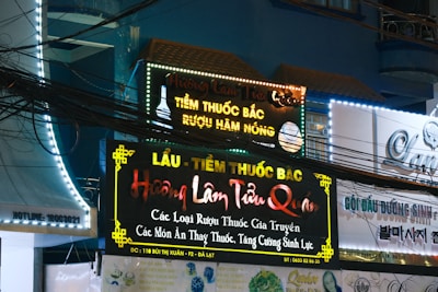 A storefront with multiple illuminated signs featuring Vietnamese text advertising traditional medicine and herbal products. The signs are colorful with bright neon lights, and there are visible electrical wires crisscrossing in front. The surrounding architecture is urban with a visible balcony and windows.