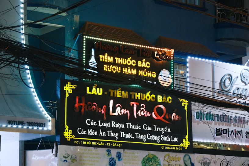 A storefront with multiple illuminated signs featuring Vietnamese text advertising traditional medicine and herbal products. The signs are colorful with bright neon lights, and there are visible electrical wires crisscrossing in front. The surrounding architecture is urban with a visible balcony and windows.