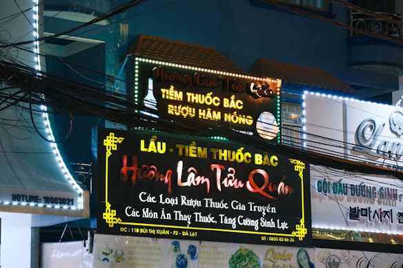A storefront with multiple illuminated signs featuring Vietnamese text advertising traditional medicine and herbal products. The signs are colorful with bright neon lights, and there are visible electrical wires crisscrossing in front. The surrounding architecture is urban with a visible balcony and windows.