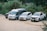 Four vehicles are parked on a dirt patch surrounded by lush greenery. The vehicles include a black SUV, a white transit van, a white SUV, and a white pickup truck. Each vehicle has decorative red bows on the front, and the setting appears to be in a remote or rural area.