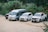 Four vehicles are parked on a dirt patch surrounded by lush greenery. The vehicles include a black SUV, a white transit van, a white SUV, and a white pickup truck. Each vehicle has decorative red bows on the front, and the setting appears to be in a remote or rural area.