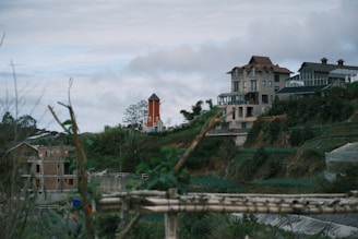 A large, multi-story house with a distinctive architectural design is situated on a hillside surrounded by greenery. A small orange windmill stands nearby, adding a quaint touch to the landscape. The cloudy sky creates a serene backdrop, with some wooden structures and construction visible in the foreground.