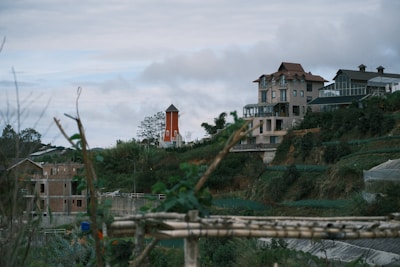 A large, multi-story house with a distinctive architectural design is situated on a hillside surrounded by greenery. A small orange windmill stands nearby, adding a quaint touch to the landscape. The cloudy sky creates a serene backdrop, with some wooden structures and construction visible in the foreground.