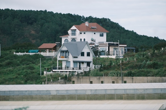 Houses situated on a green hillside with a forest in the background. The houses have modern architecture, with white walls and different roof styles. There are also some structures like greenhouses or large tents in the foreground of the image.