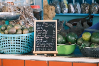 Baskets of green fruits, likely avocados, are placed on a counter. A small blackboard menu with white chalk writing stands prominently, listing various beverages and prices. Behind the counter, there are packages of dried goods.