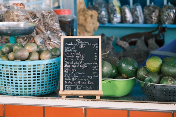 Baskets of green fruits, likely avocados, are placed on a counter. A small blackboard menu with white chalk writing stands prominently, listing various beverages and prices. Behind the counter, there are packages of dried goods.