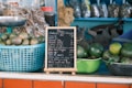 Baskets of green fruits, likely avocados, are placed on a counter. A small blackboard menu with white chalk writing stands prominently, listing various beverages and prices. Behind the counter, there are packages of dried goods.