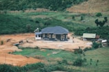 A small house with a dark gray roof is situated on a cleared patch of land surrounded by lush greenery and farmland. The area around the house appears to be under development, with various construction materials and machinery scattered around. An adjacent smaller structure with a green roof is also visible.