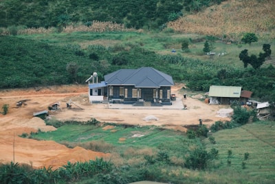 A small house with a dark gray roof is situated on a cleared patch of land surrounded by lush greenery and farmland. The area around the house appears to be under development, with various construction materials and machinery scattered around. An adjacent smaller structure with a green roof is also visible.
