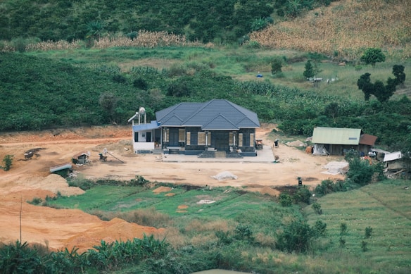 A small house with a dark gray roof is situated on a cleared patch of land surrounded by lush greenery and farmland. The area around the house appears to be under development, with various construction materials and machinery scattered around. An adjacent smaller structure with a green roof is also visible.