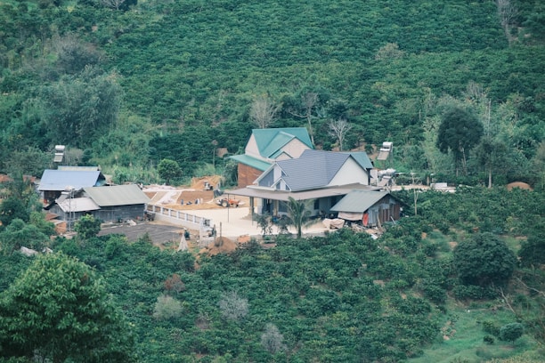 A rural farm surrounded by lush green vegetation, featuring several buildings with metal roofs, some equipment, and open spaces. The setting is amidst a dense, forested area on rolling hills.