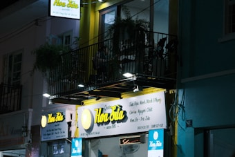 A nighttime street scene shows a small business establishment with a brightly lit sign that reads 'Hoa Sữa' and offers various items such as coffee and soymilk. Above the shop, there is a balcony with plants and furniture, and the ambience is calm and quiet.