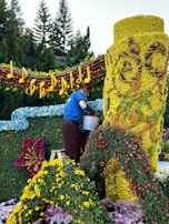 A person is arranging flowers on a large floral display. The display features a bright yellow cylindrical structure covered in flowers, intricate designs, and surrounded by a variety of colorful blossoms. Musical notes and a staff made of flowers and foliage adorn the top, while trees can be seen in the background.