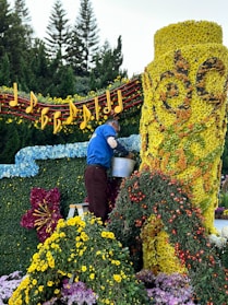 A person is arranging flowers on a large floral display. The display features a bright yellow cylindrical structure covered in flowers, intricate designs, and surrounded by a variety of colorful blossoms. Musical notes and a staff made of flowers and foliage adorn the top, while trees can be seen in the background.