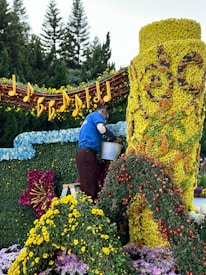 A person is arranging flowers on a large floral display. The display features a bright yellow cylindrical structure covered in flowers, intricate designs, and surrounded by a variety of colorful blossoms. Musical notes and a staff made of flowers and foliage adorn the top, while trees can be seen in the background.