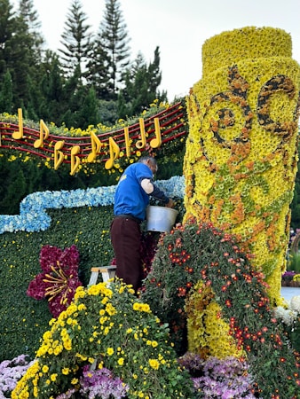 A person is arranging flowers on a large floral display. The display features a bright yellow cylindrical structure covered in flowers, intricate designs, and surrounded by a variety of colorful blossoms. Musical notes and a staff made of flowers and foliage adorn the top, while trees can be seen in the background.