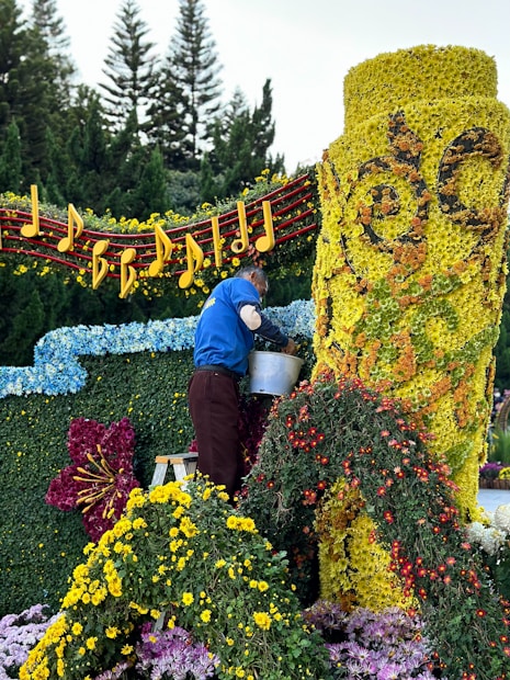 A person is arranging flowers on a large floral display. The display features a bright yellow cylindrical structure covered in flowers, intricate designs, and surrounded by a variety of colorful blossoms. Musical notes and a staff made of flowers and foliage adorn the top, while trees can be seen in the background.