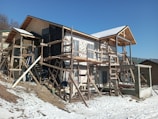 A construction site of a house being built with wooden scaffolding surrounding the structure. The ground is covered with snow, and the sky is clear with a bright blue hue. The house is in the early stages of construction with a visible framework.