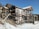 A construction site of a house being built with wooden scaffolding surrounding the structure. The ground is covered with snow, and the sky is clear with a bright blue hue. The house is in the early stages of construction with a visible framework.
