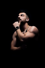 A muscular man is seen in a contemplative pose, with his hand placed under his chin. He is wearing a smartwatch and wireless earbuds, and a necklace is visible around his neck. The lighting creates a dramatic effect, highlighting his facial features and well-defined arms against a dark background.