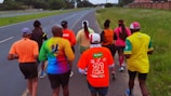A group of people are jogging on a paved road that runs parallel to a grassy area under a slightly overcast sky. They are wearing colorful athletic shirts and caps, with text and logos visible on the clothing.