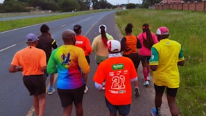 A group of people are jogging on a paved road that runs parallel to a grassy area under a slightly overcast sky. They are wearing colorful athletic shirts and caps, with text and logos visible on the clothing.