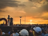 A singer stands on a stage holding a guitar in front of a large crowd of people. The backdrop is a sunset sky with warm hues and scattered clouds. String lights hang above the crowd, adding a festive atmosphere. The audience is gathered closely, and the scene is lively and engaging.