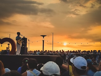 A singer stands on a stage holding a guitar in front of a large crowd of people. The backdrop is a sunset sky with warm hues and scattered clouds. String lights hang above the crowd, adding a festive atmosphere. The audience is gathered closely, and the scene is lively and engaging.