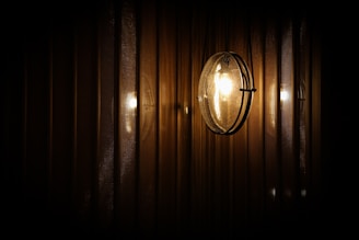 A spherical wall-mounted light fixture emits a warm glow, reflecting against a series of vertical, textured metallic or wooden panels. The lighting creates a subtle play of shadows and highlights, giving a cozy and intimate ambiance.