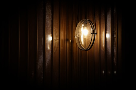 A spherical wall-mounted light fixture emits a warm glow, reflecting against a series of vertical, textured metallic or wooden panels. The lighting creates a subtle play of shadows and highlights, giving a cozy and intimate ambiance.