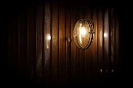 A spherical wall-mounted light fixture emits a warm glow, reflecting against a series of vertical, textured metallic or wooden panels. The lighting creates a subtle play of shadows and highlights, giving a cozy and intimate ambiance.