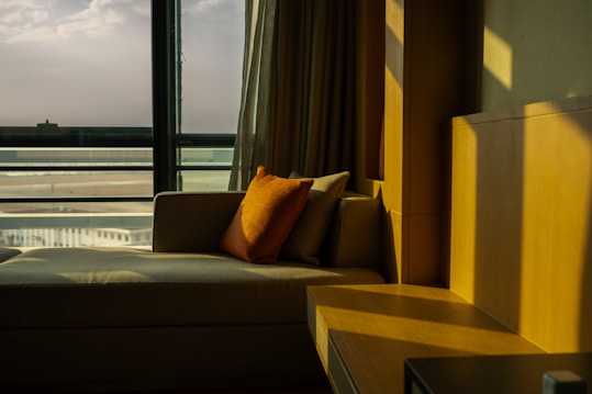 A cozy indoor setting with a plush beige sofa positioned next to a large window. An orange pillow rests on the sofa, adding a pop of color. Soft light streams in through the window, casting warm shadows on the wooden furniture and walls. Outside, a distant view of a beach and horizon can be seen through the glass.