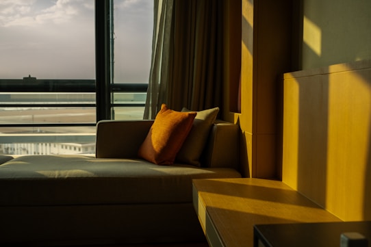 A cozy indoor setting with a plush beige sofa positioned next to a large window. An orange pillow rests on the sofa, adding a pop of color. Soft light streams in through the window, casting warm shadows on the wooden furniture and walls. Outside, a distant view of a beach and horizon can be seen through the glass.
