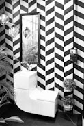 A modern, monochrome bathroom corner featuring a sleek white combination unit with a washbasin and toilet. The wall presents a bold black and white chevron pattern. Above the unit is a rectangular mirror reflecting hanging glass pendant lights. To the right is a small tiered stand holding round potted plants. A tall leafy plant peeks into the frame on the left side.