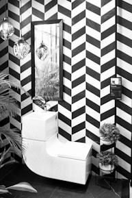 A modern, monochrome bathroom corner featuring a sleek white combination unit with a washbasin and toilet. The wall presents a bold black and white chevron pattern. Above the unit is a rectangular mirror reflecting hanging glass pendant lights. To the right is a small tiered stand holding round potted plants. A tall leafy plant peeks into the frame on the left side.