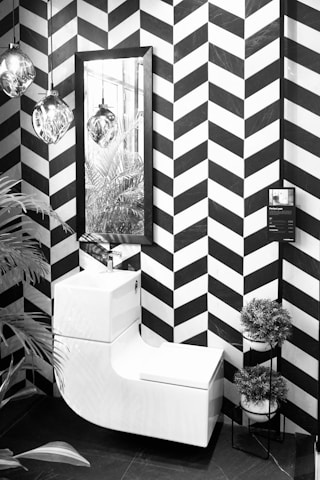 A modern, monochrome bathroom corner featuring a sleek white combination unit with a washbasin and toilet. The wall presents a bold black and white chevron pattern. Above the unit is a rectangular mirror reflecting hanging glass pendant lights. To the right is a small tiered stand holding round potted plants. A tall leafy plant peeks into the frame on the left side.