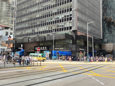 A busy urban street corner featuring a tall office building with reflective glass windows and various bank logos. Pedestrians are crossing the street and waiting at the intersection. A yellow and green minibus is passing by, and there are advertisements and signs in both English and Chinese.