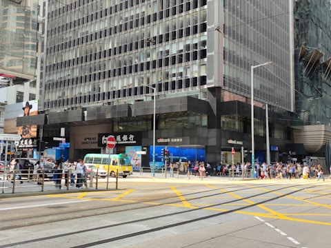 A busy urban street corner featuring a tall office building with reflective glass windows and various bank logos. Pedestrians are crossing the street and waiting at the intersection. A yellow and green minibus is passing by, and there are advertisements and signs in both English and Chinese.