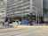 A busy urban street corner featuring a tall office building with reflective glass windows and various bank logos. Pedestrians are crossing the street and waiting at the intersection. A yellow and green minibus is passing by, and there are advertisements and signs in both English and Chinese.