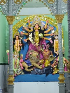 A detailed sculpture of a multi-armed goddess standing on a platform, surrounded by attendants and a lion. The goddess is adorned with elaborate jewelry and colorful robes, holding various weapons. The ornate background features intricate patterns and bright colors.