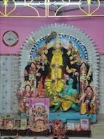A richly decorated Hindu goddess Durga sculpture is depicted, with multiple arms holding various objects. The figure is adorned with vibrant garlands and jewelry, set against a colorful and ornate backdrop. Additional figures and religious items are arranged around the sculpture, including images and small decorative elements. A clock and decorative panel are visible in the background.
