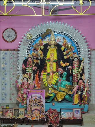 A richly decorated Hindu goddess Durga sculpture is depicted, with multiple arms holding various objects. The figure is adorned with vibrant garlands and jewelry, set against a colorful and ornate backdrop. Additional figures and religious items are arranged around the sculpture, including images and small decorative elements. A clock and decorative panel are visible in the background.