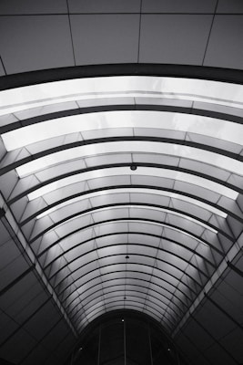 An architectural ceiling with a series of curved, parallel beams forming a tunnel-like structure. The design is symmetric and creates a play of light and shadows with a minimalist style.