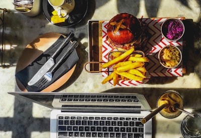 A top-down view of a table with a meal and a laptop. A burger with fries is served on a tray with chevron-patterned paper, accompanied by small bowls of purple cabbage salad and coleslaw. A laptop is open nearby, and a drink with a straw is placed to the side. Utensils rest on a napkin on a plate.