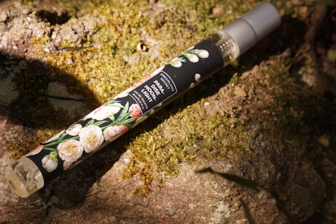 A slim perfume bottle with a floral design label and gray cap rests on a mossy, sunlit stone surface. The label reads 'PARADISE MOONLIGHT' with additional text indicating it's an eau de parfum.