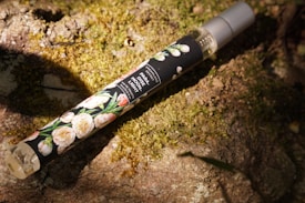 A slim perfume bottle with a floral design label and gray cap rests on a mossy, sunlit stone surface. The label reads 'PARADISE MOONLIGHT' with additional text indicating it's an eau de parfum.