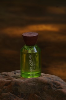 A green perfume bottle with a wooden cap placed on a textured rock surface. The background is blurred with warm amber lighting, creating a natural and earthy ambiance.