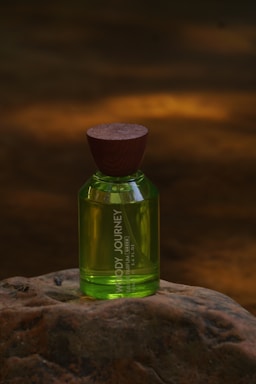 A green perfume bottle with a wooden cap placed on a textured rock surface. The background is blurred with warm amber lighting, creating a natural and earthy ambiance.